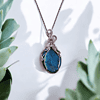 Bohemian wire-wrapped pendant with blue healing crystal and vintage-style wire, surrounded by green succulent leaves.