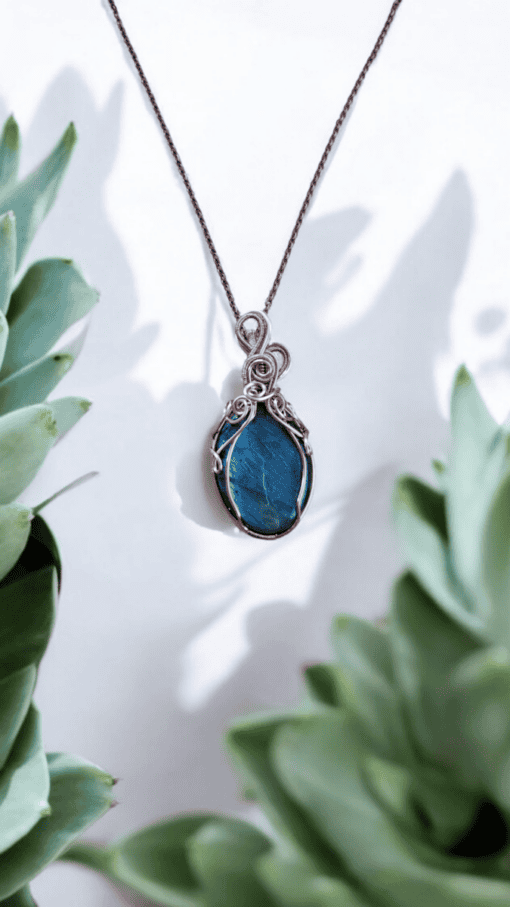 Bohemian wire-wrapped pendant with blue healing crystal and vintage-style wire, surrounded by green succulent leaves.