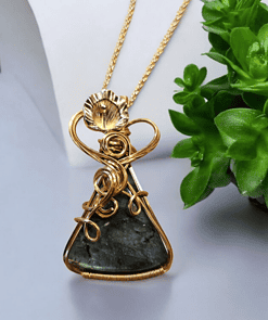 Golden pendant with intricate wirework and a dark triangular stone on a chain, placed beside a small green plant.