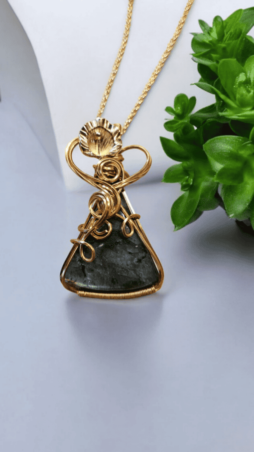 Golden pendant with intricate wirework and a dark triangular stone on a chain, placed beside a small green plant.