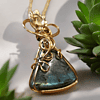 Gold wire-wrapped blue gemstone pendant on a gold chain, surrounded by green succulent plants.