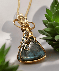 Gold wire-wrapped blue gemstone pendant on a gold chain, surrounded by green succulent plants.