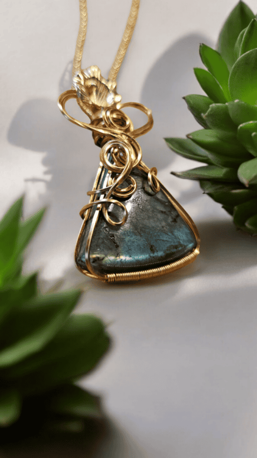 Gold wire-wrapped blue gemstone pendant on a gold chain, surrounded by green succulent plants.