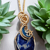 Gold-wire wrapped blue stone pendant with a crescent moon and star design, placed between two green succulent plants.