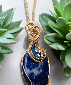 Gold-wire wrapped blue stone pendant with a crescent moon and star design, placed between two green succulent plants.