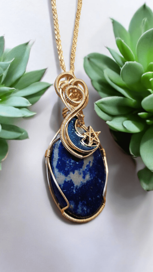 Gold-wire wrapped blue stone pendant with a crescent moon and star design, placed between two green succulent plants.