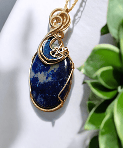 Blue stone pendant with gold wire wrapping, featuring a crescent moon and a star charm, placed on a white background with green leaves.