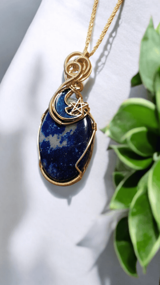 Blue stone pendant with gold wire wrapping, featuring a crescent moon and a star charm, placed on a white background with green leaves.