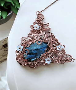 Ornate copper wire-wrapped pendant with a blue stone and floral accents, displayed on a white background with green plants nearby.