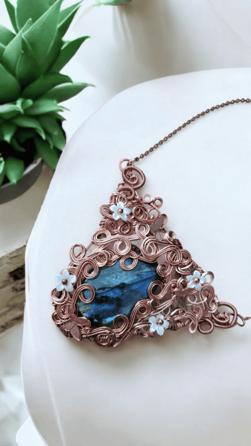 Ornate copper wire-wrapped pendant with a blue stone and floral accents, displayed on a white background with green plants nearby.