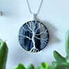 A round pendant necklace with a silver wire tree design on a dark blue stone background, surrounded by green succulents.