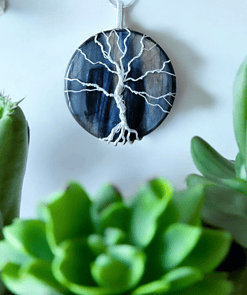 A round pendant necklace with a silver wire tree design on a dark blue stone background, surrounded by green succulents.