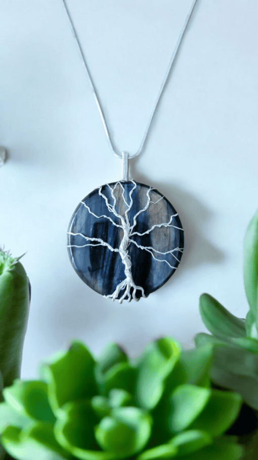 A round pendant necklace with a silver wire tree design on a dark blue stone background, surrounded by green succulents.