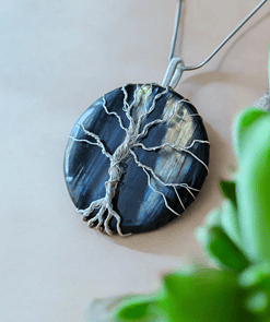 Round pendant with wire-wrapped tree design on dark stone background, laying on beige surface with green plant near it.