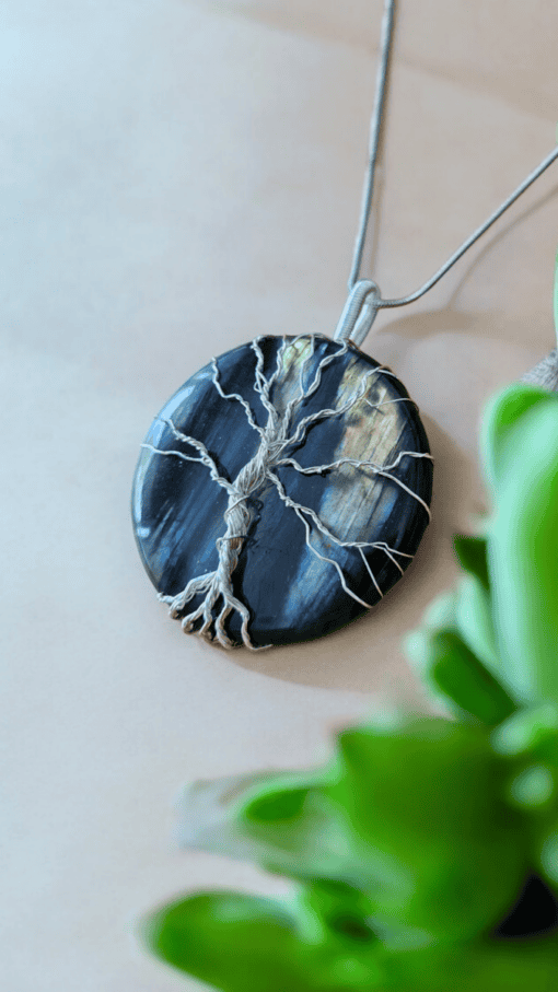 Round pendant with wire-wrapped tree design on dark stone background, laying on beige surface with green plant near it.