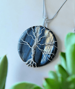 A round pendant with a tree design made of wire on a dark blue stone, with green leaves in the foreground.