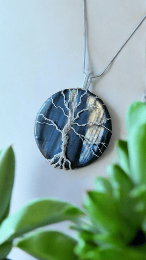 A round pendant with a tree design made of wire on a dark blue stone, with green leaves in the foreground.