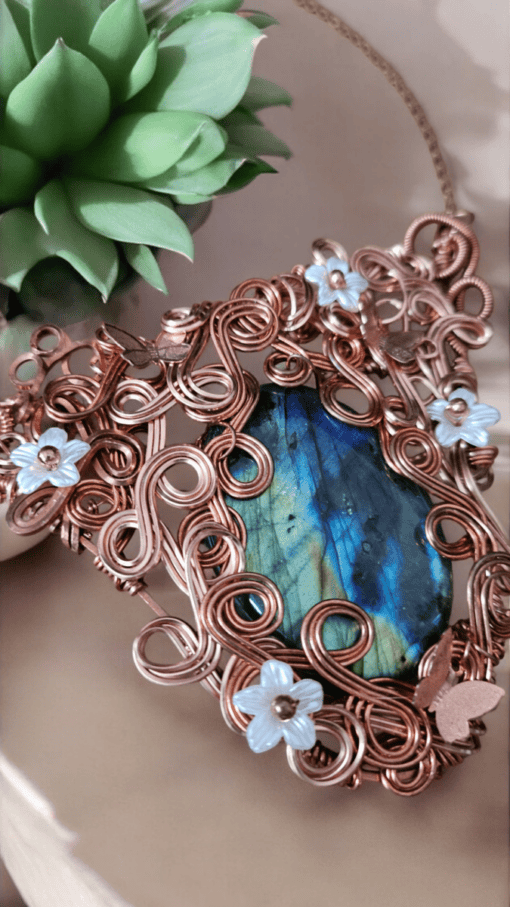 Copper wire pendant with a blue-green gemstone, decorated with light blue and copper flowers, next to a small green succulent.