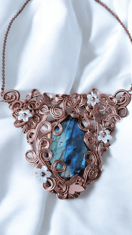 Copper wire necklace with blue gemstone, intricate swirls, butterflies, and flower designs, displayed on white fabric.