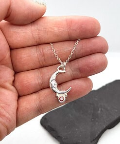 Hand holding a silver crescent moon necklace with a small orange gemstone at the bottom, shown against a black background.