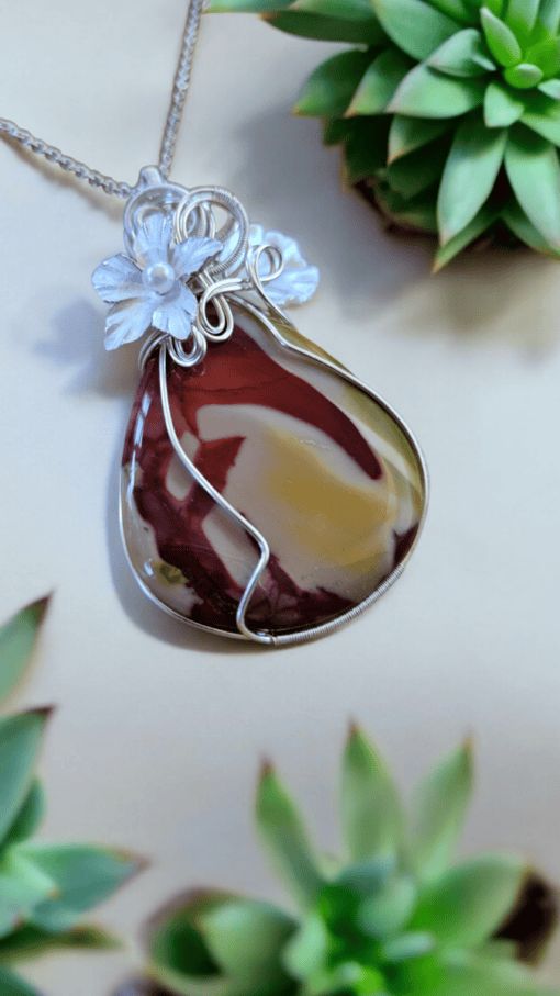 Handmade wire wrapped pendant with floral accents and multicolored stone, placed near green succulents. Unique and artisan design.