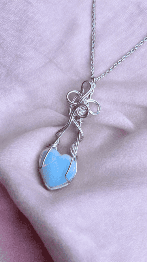 Handcrafted heart jewellery featuring a delicate blue heart charm necklace with intricate wire wrapping, on a soft pink background.