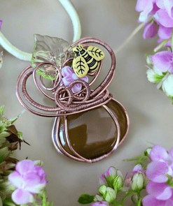 Handmade pendant with wire wrapping, a brown stone, a glass leaf, and a small bee charm, surrounded by pink flowers.