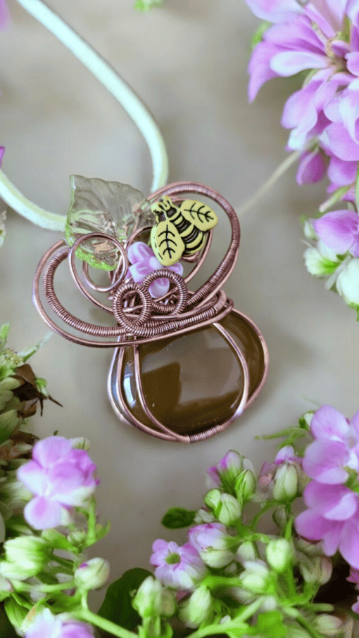 Handmade pendant with wire wrapping, a brown stone, a glass leaf, and a small bee charm, surrounded by pink flowers.