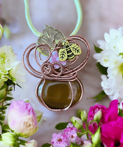 Handmade necklace with green pendant, wire wrapping, and bee charm, surrounded by pink and white flowers.