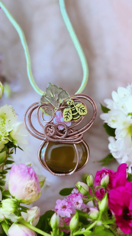 Handmade necklace with green pendant, wire wrapping, and bee charm, surrounded by pink and white flowers.