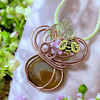 A pendant featuring a brown stone, wire loops, a small flower, and a bee charm on a green cord, surrounded by green and pink flowers.