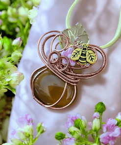 A pendant featuring a brown stone, wire loops, a small flower, and a bee charm on a green cord, surrounded by green and pink flowers.