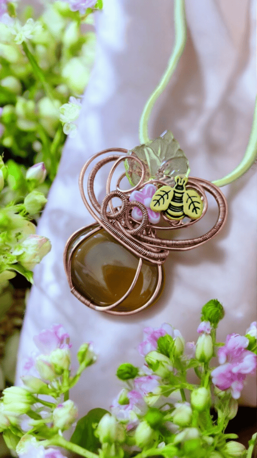 A pendant featuring a brown stone, wire loops, a small flower, and a bee charm on a green cord, surrounded by green and pink flowers.