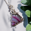 Silver wire-wrapped pendant with a purple striped triangular gemstone, next to green succulent leaves on a light background.