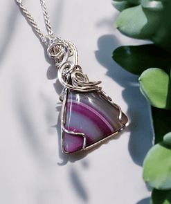 Silver wire-wrapped pendant with a purple striped triangular gemstone, next to green succulent leaves on a light background.