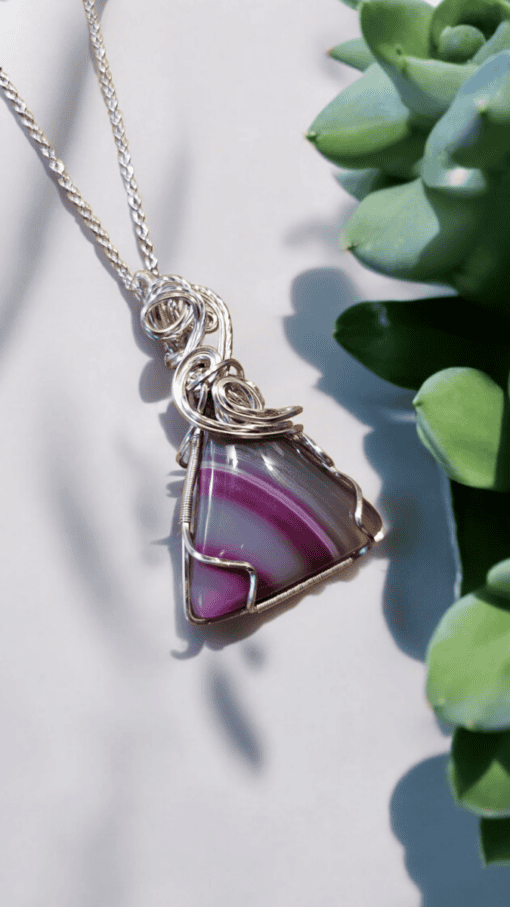 Silver wire-wrapped pendant with a purple striped triangular gemstone, next to green succulent leaves on a light background.