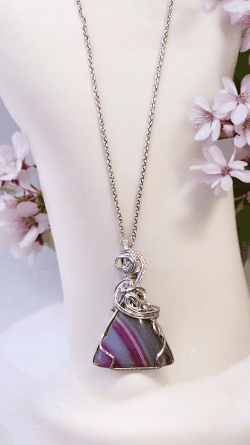 Silver wire-wrapped necklace with a triangular purple and grey pendant, displayed on a white bust with pink flowers.