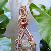 Pendant with a copper wire-wrapped gemstone, hanging against a backdrop of green leaves.