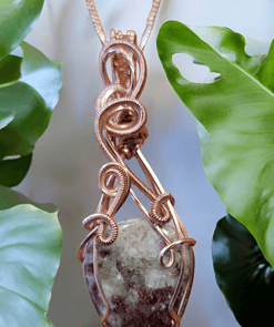 Pendant with a copper wire-wrapped gemstone, hanging against a backdrop of green leaves.