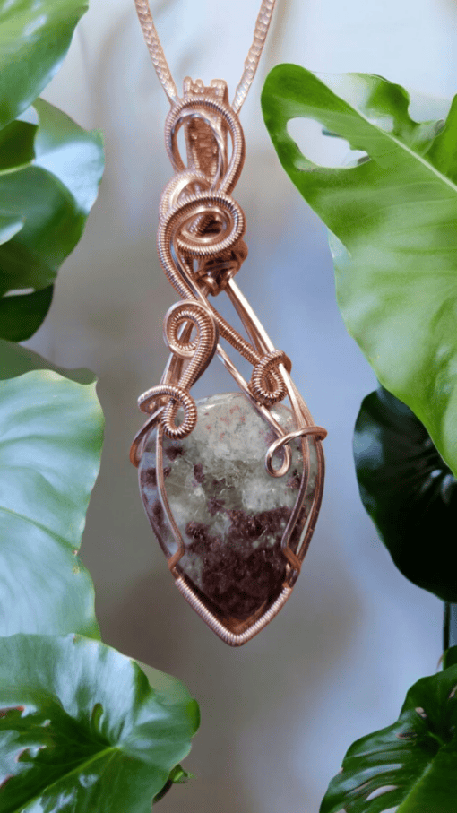 Pendant with a copper wire-wrapped gemstone, hanging against a backdrop of green leaves.