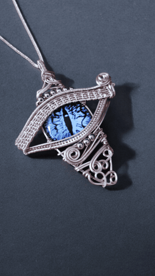 Intricate silver wire-wrapped pendant with a blue cat-eye stone at the center, resembling an eye, on a dark background.