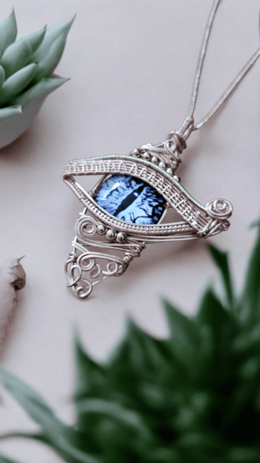 Intricate silver wire-wrapped pendant with a blue eye design, placed among green succulents on a light background.