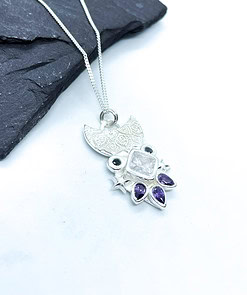 Silver owl-shaped necklace with detailed patterns, featuring purple and clear gemstones, placed on a dark slate background.