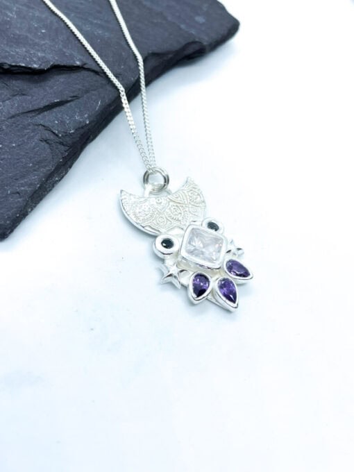 Silver owl-shaped necklace with detailed patterns, featuring purple and clear gemstones, placed on a dark slate background.