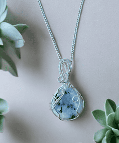 Silver wire-wrapped pendant with a blue and black stone, surrounded by green succulents on a beige background.