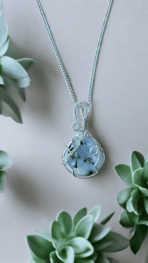 Silver wire-wrapped pendant with a blue and black stone, surrounded by green succulents on a beige background.