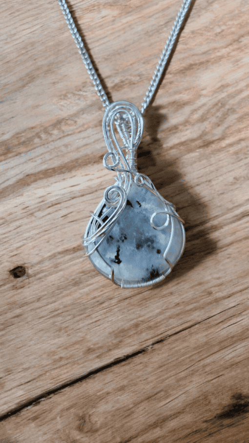 Silver wire-wrapped pendant with a translucent stone featuring black inclusions, displayed on a wooden surface.