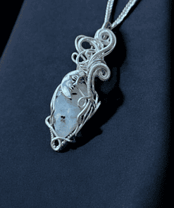 Silver wire-wrapped necklace with an ornate moon face and clear stone pendant on a dark blue fabric background.