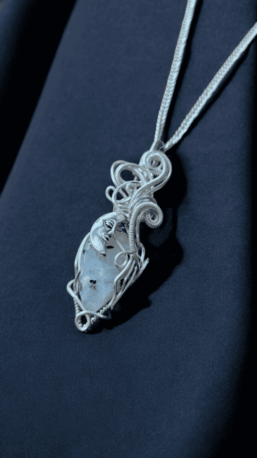 Silver wire-wrapped necklace with an ornate moon face and clear stone pendant on a dark blue fabric background.