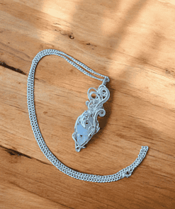 Silver pendant with intricate wire wrapping on a chain, displayed on a wooden surface with sunlight casting shadows.